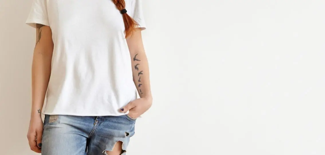 Cropped shot of beautiful stylish young redhead woman with braid and tattoos smiling joyfully dressed in white oversize t-shirt, keeping hand in pocket of her trendy blue ragged jeans.