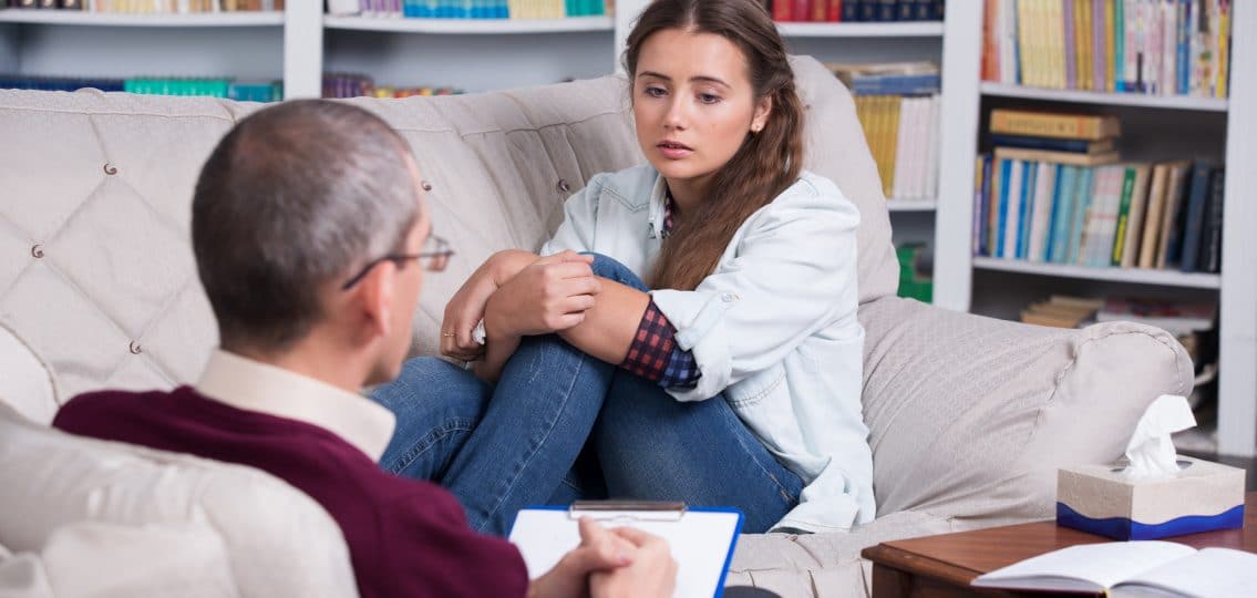 Teenage girl crying on sofa during therapy session while therapist is taking notes