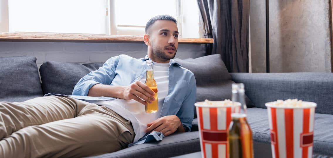 college kid drinking at home on the couch watching tv holding a beer