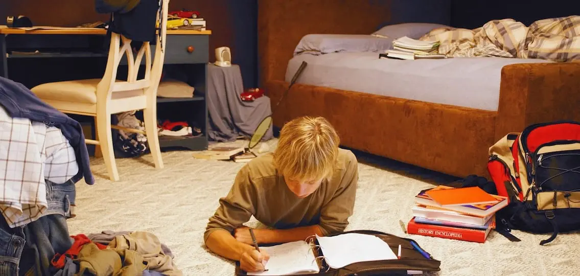 Boy Doing Homework In Messy Room surrounded by homework and clothes