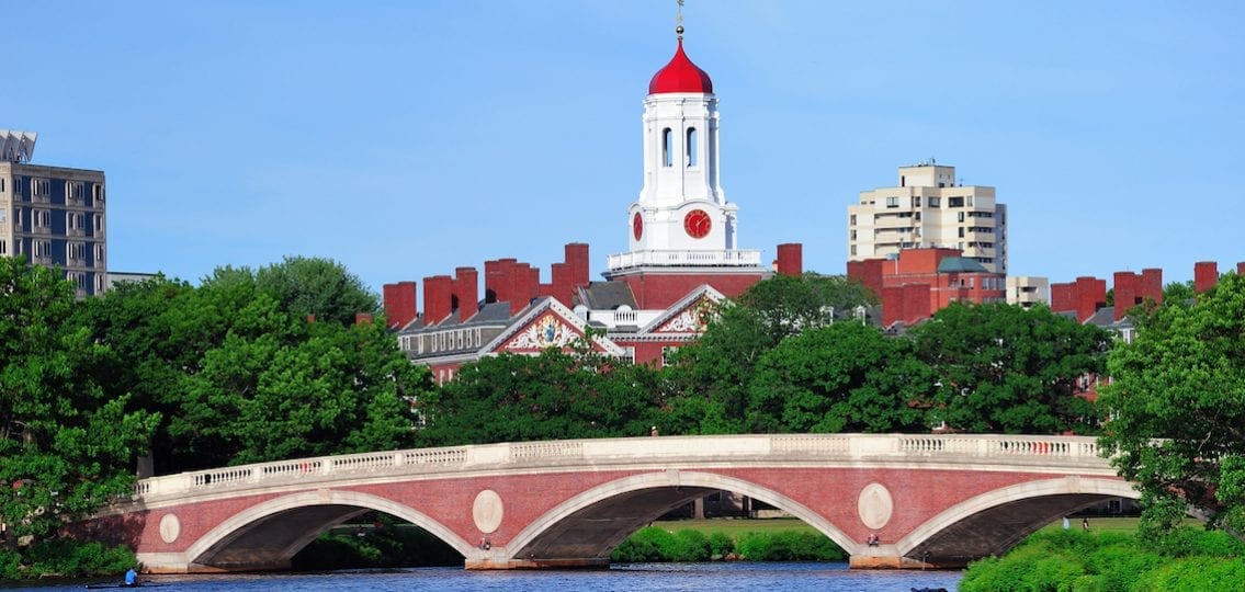 John W. Weeks Bridge and clock tower over Charles River in Harvard University campus in Boston with trees, boat and blue sky.