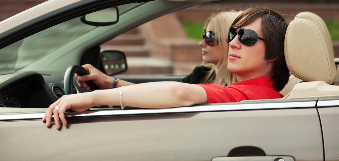 Happy young teenage couple driving convertible car wearing sunglasses
