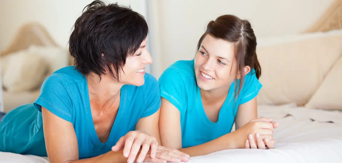 middle aged mother and teen daughter talking on bed and smiling