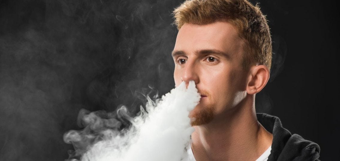 teen boy vaping and breathing out smoke on a black background