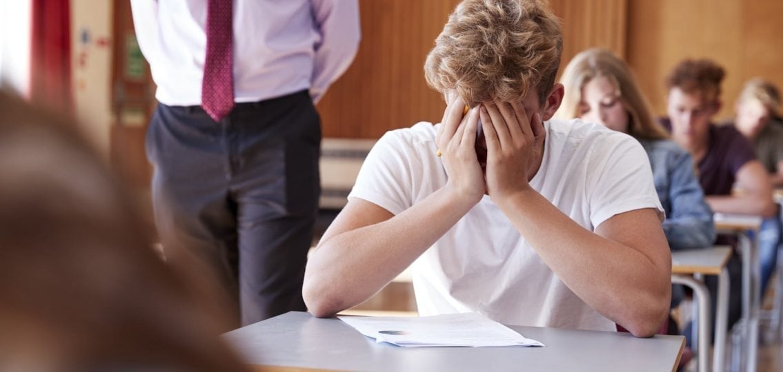 anxious teen boy with his face in his hands during a test as the teacher walks past