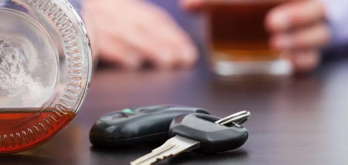 car keys on the table next to a beer with a blurred figure drinking alcohol in the background