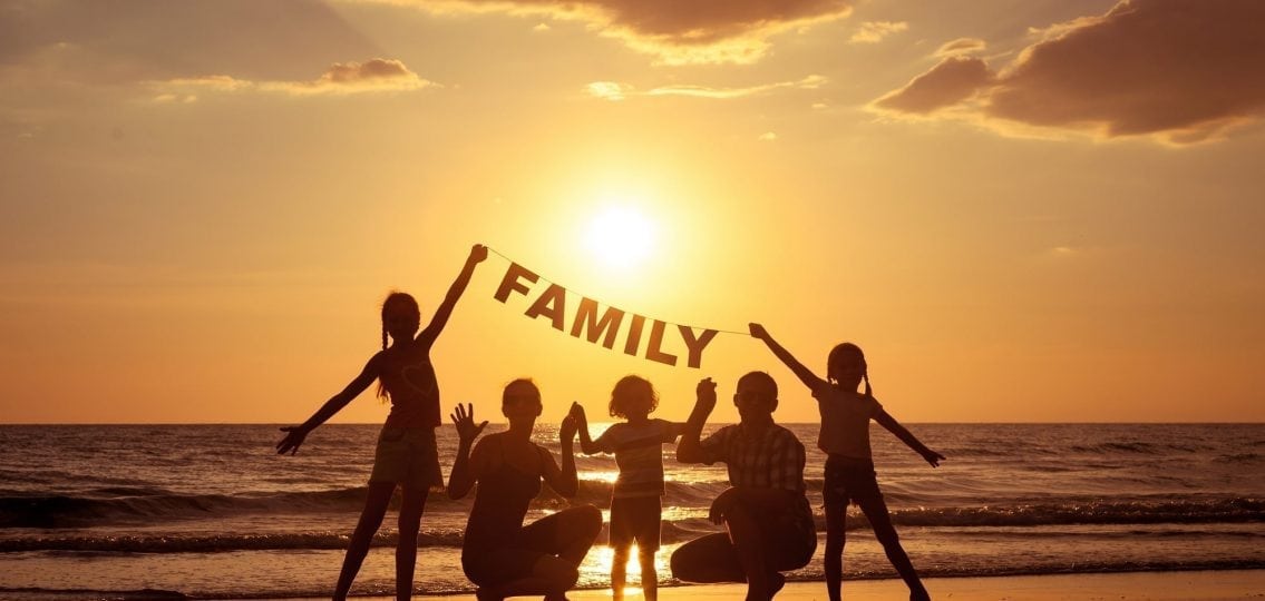 blended family on the beach silhouetted in front of the sunset holding a sign that says family