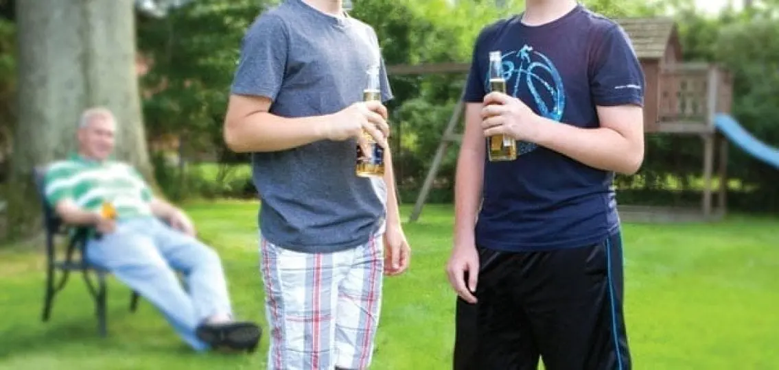 two teenagers drinking beer outside in a backyard as a dad drinks on a lawn chair in the background