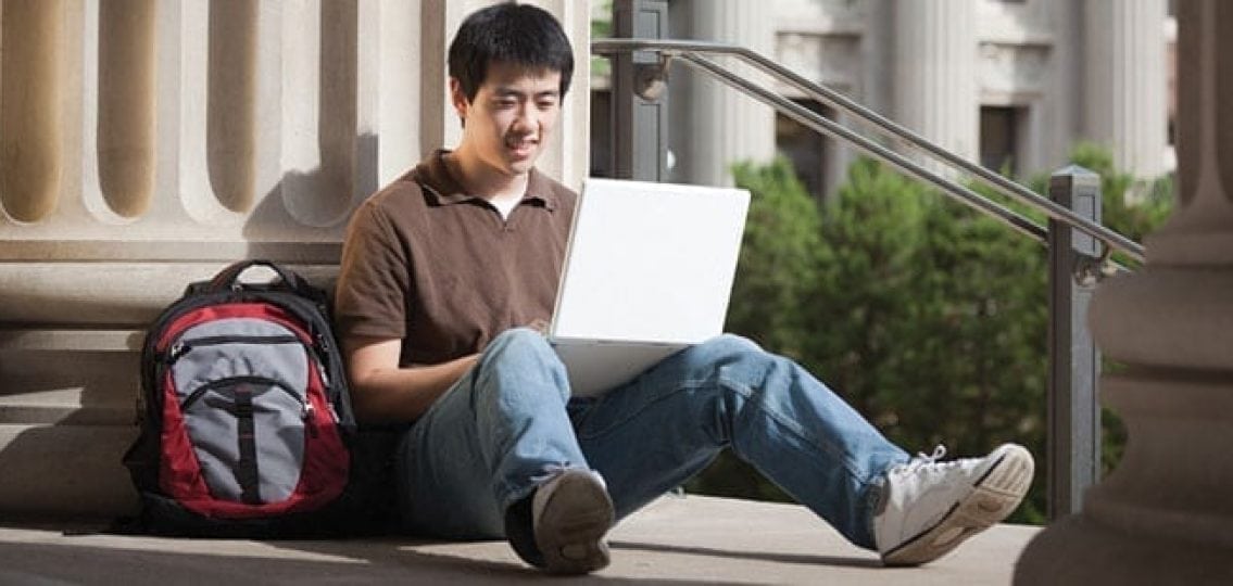 college student sitting on the steps with his backpack typing on a computer outdoors