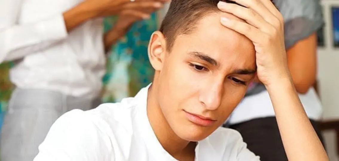 tearful anxious teenage boy grabbing head with parents in the background