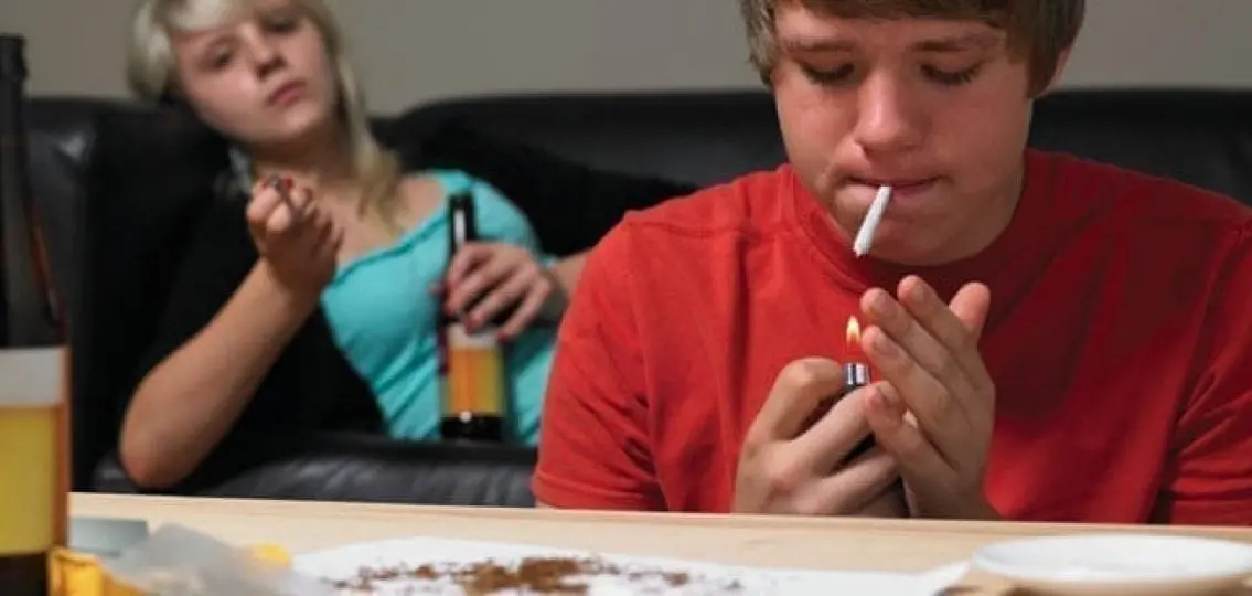 teen boy smoking a joint while a teen girl drinks beer and smokes weed in the background