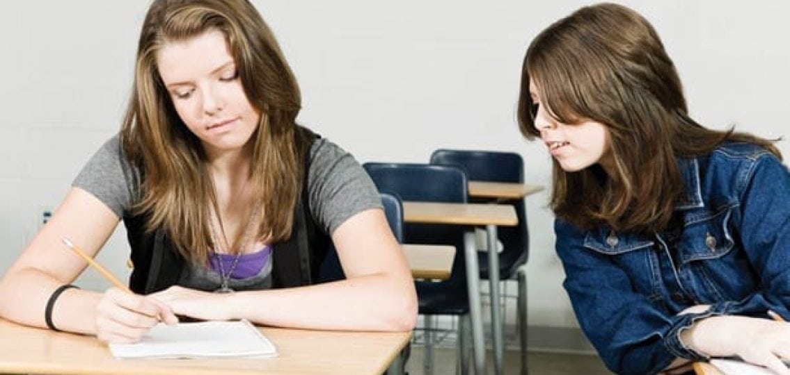 a teenage girl leaning over to cheat on her homework and copy her friend