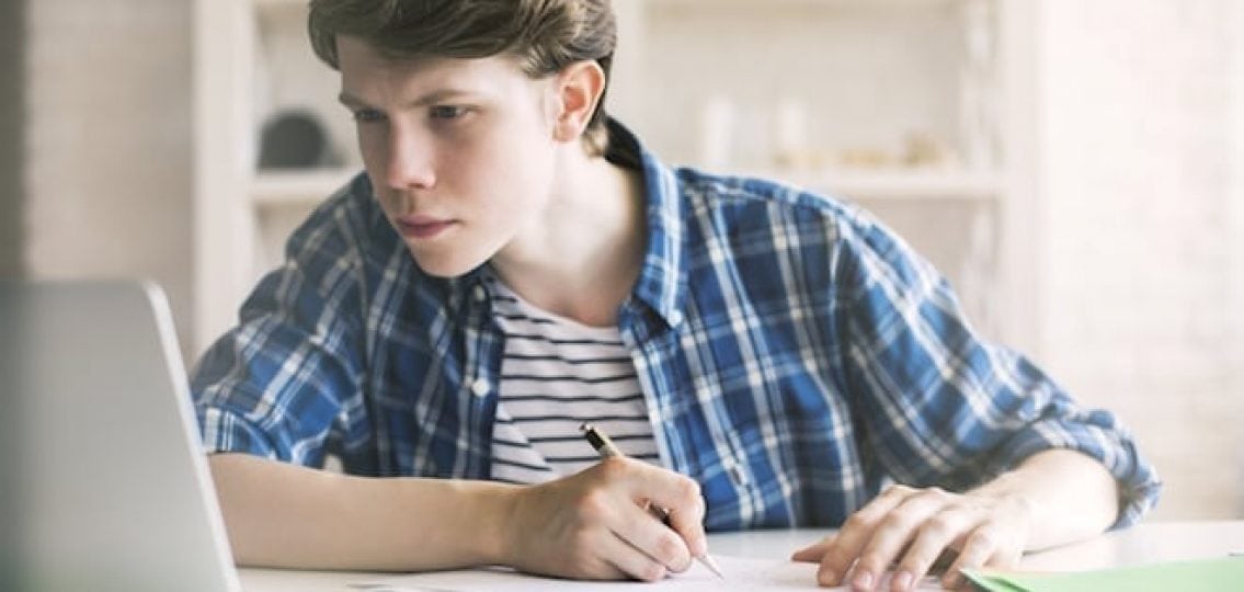 teen writing a paper with a pen looking at his laptop