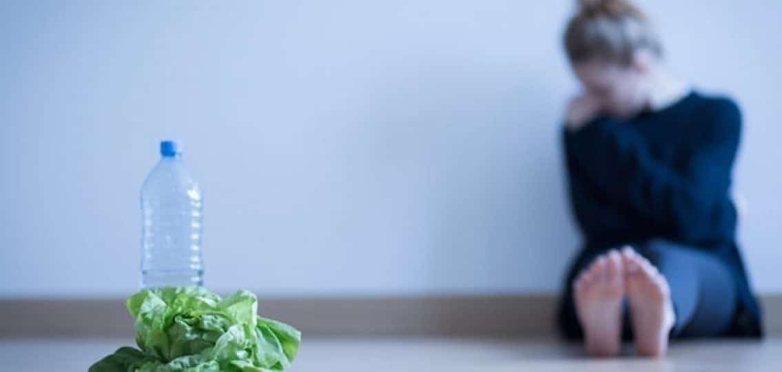 anorexic teenage girl sitting against a wall with lettuce and water bottle in foreground