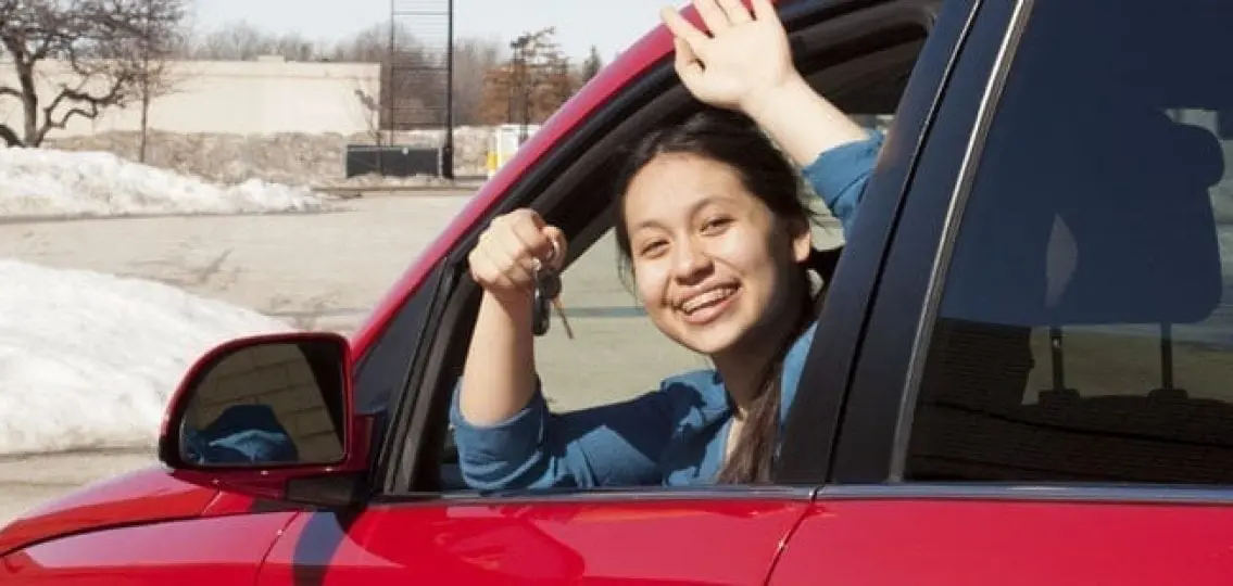 teenage girl waving out of the window while holding up her keys in a snowy parking lot