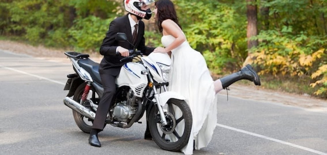 young woman in a wedding dress leaning into a man on a motorcycle in a suit eloping
