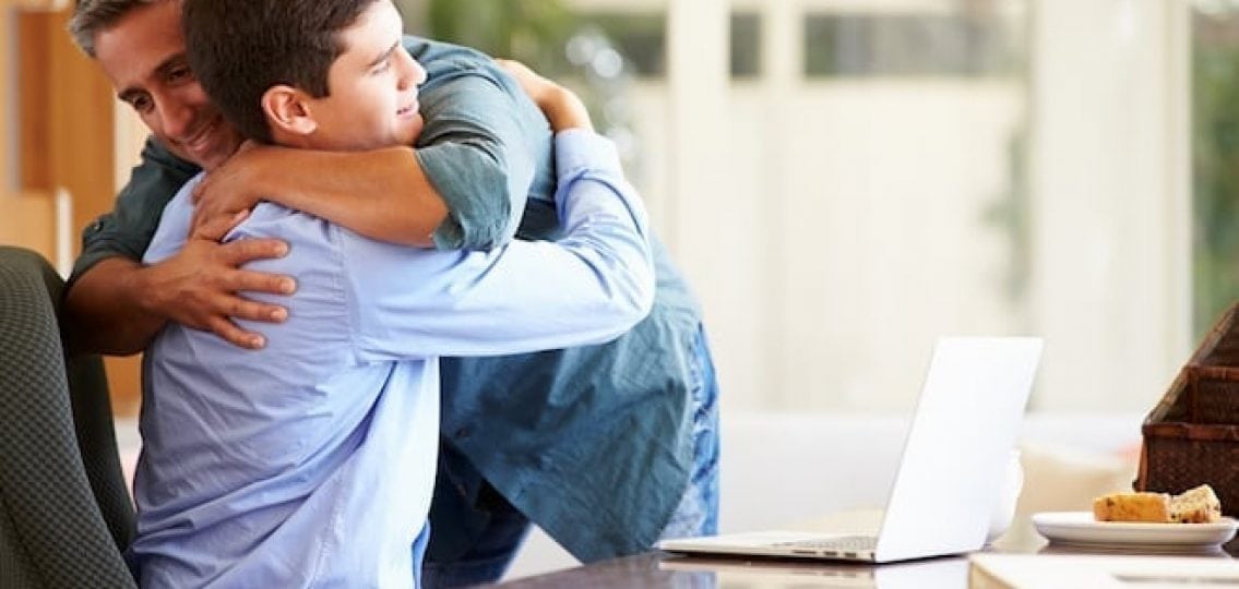 dad hugging his upset teenage son at a kitchen table in front of his laptop