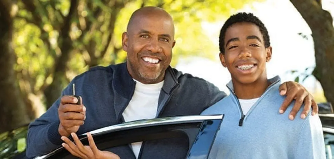 father and teenage boy with braces next to a car dad with arm around son and handing him car keys