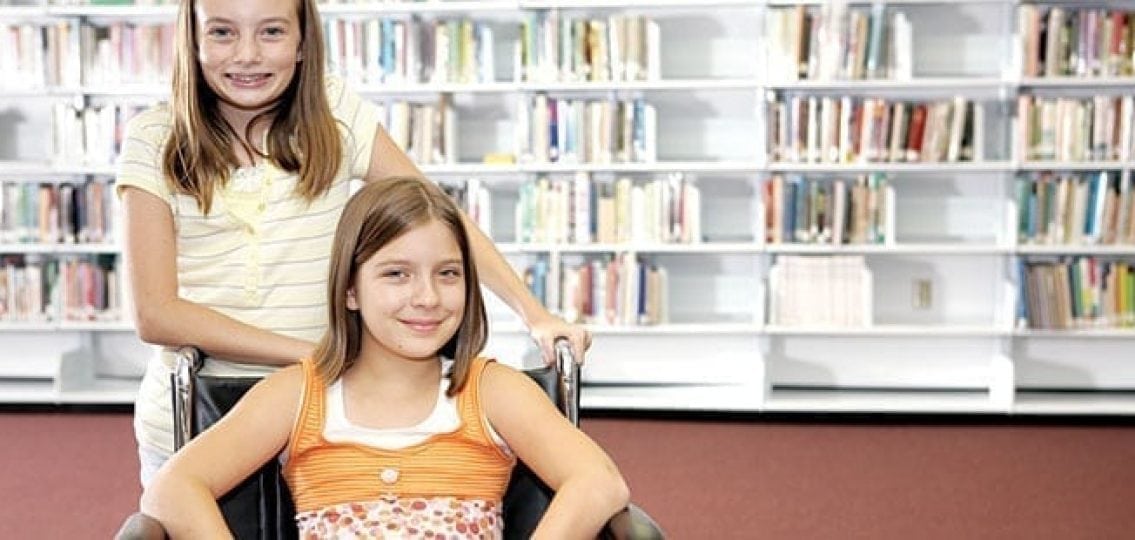 two teen girls friends in the library one girl in a wheelchair both smiling