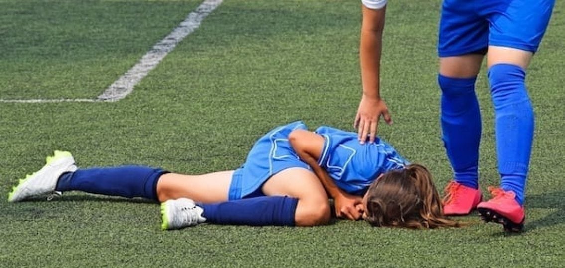 Teen girl soccer player facedown on turf with leg injury while friend checks on her