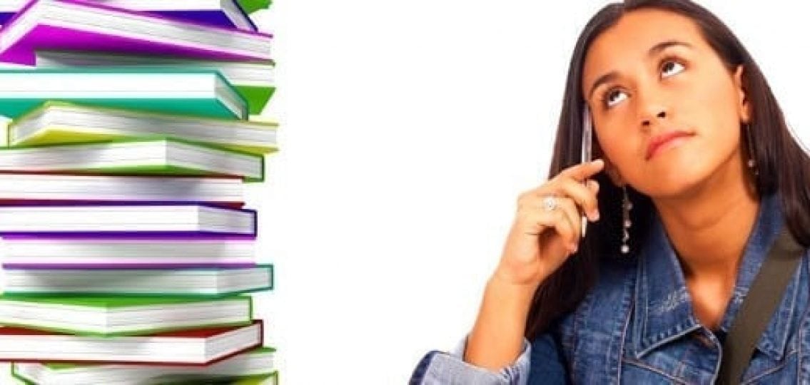 teenage girl thinking tapping a pen to her head next to a pile of textbooks stretching far over her head