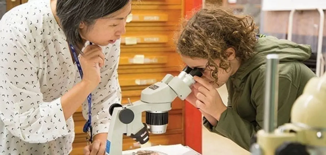 teenage girl looking in a microscope while teacher watches