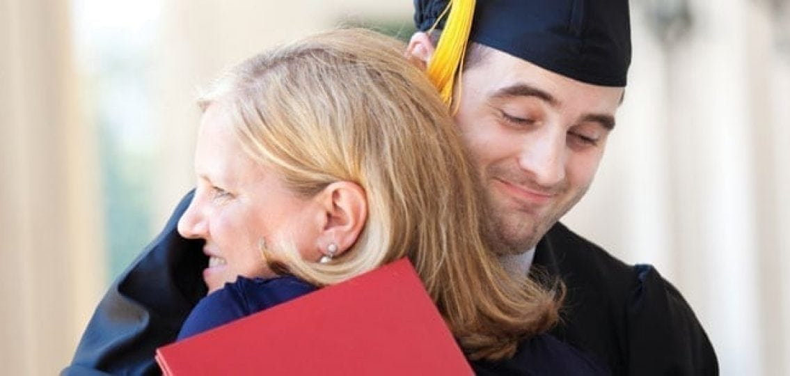 a tall teenage boy in graduation robes and a diploma hugging hit mom