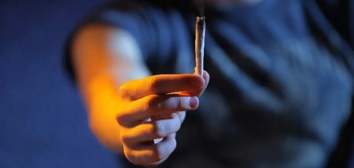 a teen girl holding out a weed joint to the camera in a dark room