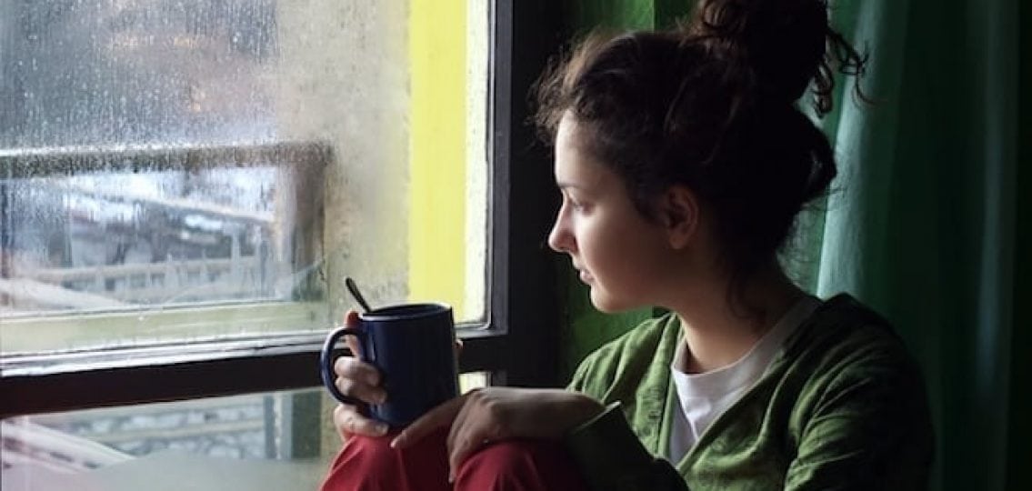 sad homesick girl looking out a rainy window with a mug of tea