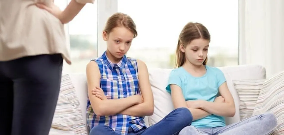 two teen sisters sitting on a couch getting scolded by their mother
