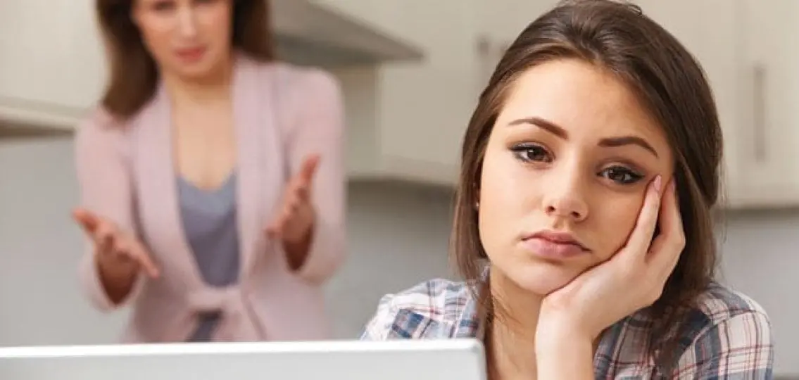 exhausted annoyed teen girl leaning on her hand while mom gets mad in the background
