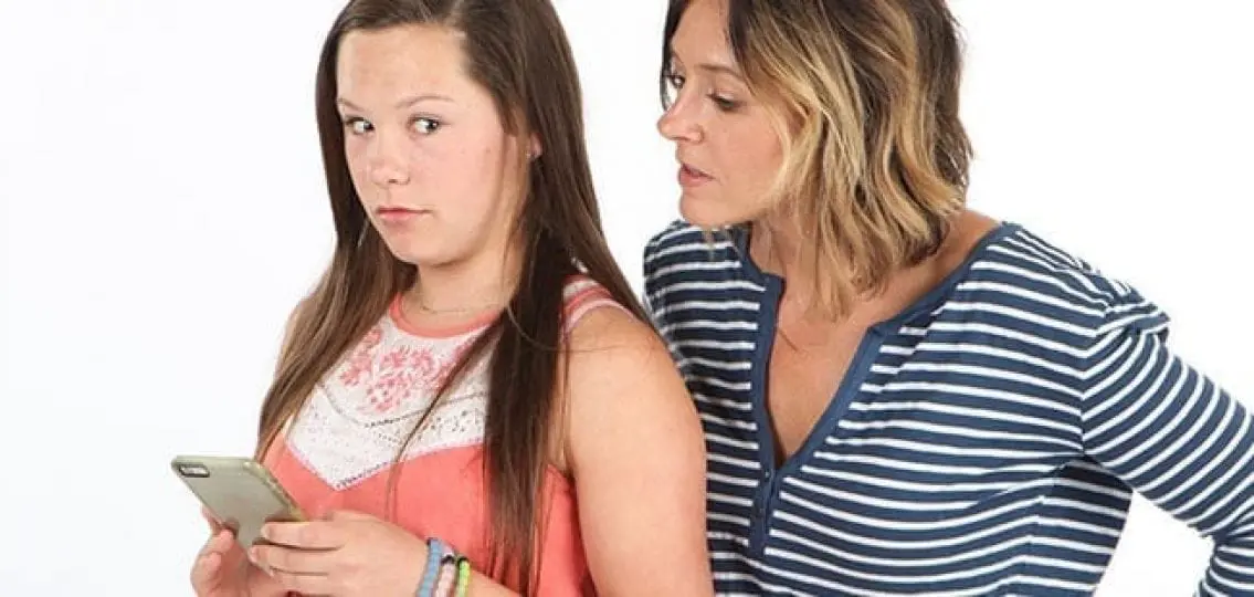 mom looking at daughter's phone over her shoulder while her daughter looks uncomfortable