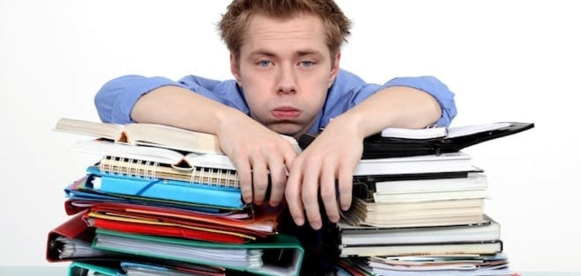 overwhelmed teenage boy leaning on piles of textbooks notebooks and binders