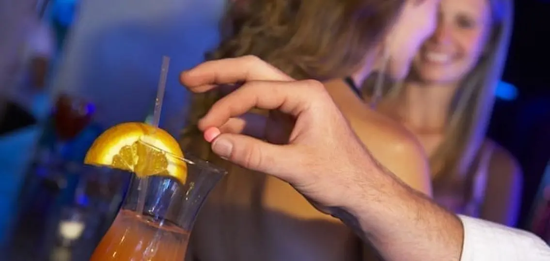 a man's hand putting a roofie into an alcoholic drink as two blurred college girls talk in the background