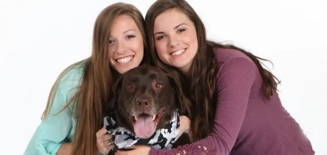 two teen sisters hugging their chocolate lab dog in a gray scarf