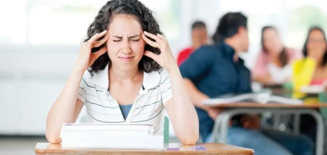 teenage girl with a headache sitting at a desk with other students blurred in background