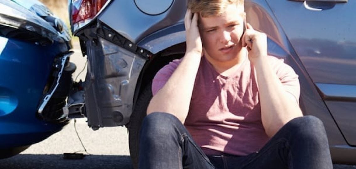 teenage boy sitting next to his crashed car blue and silver car fender bender