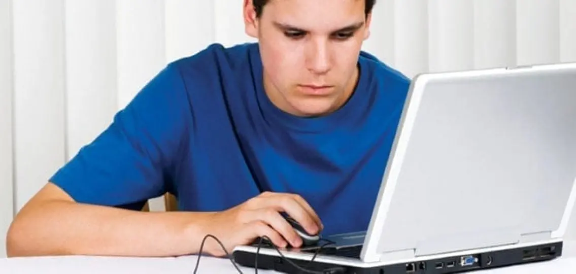 teenage boy frowning while on a laptop in front of some closed blinds