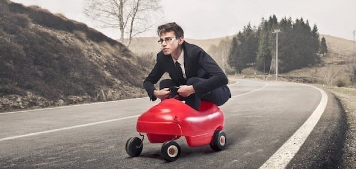 teenage boy in a suit in a tiny toy car driving down the street
