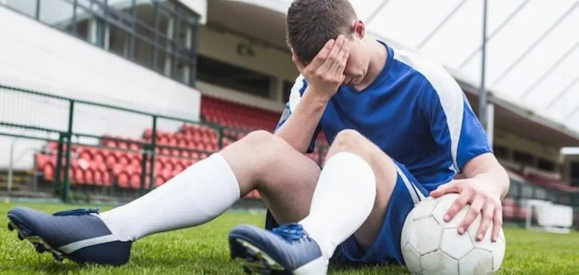 Upset teen soccer player with face in hand sitting on turf with a soccer ball