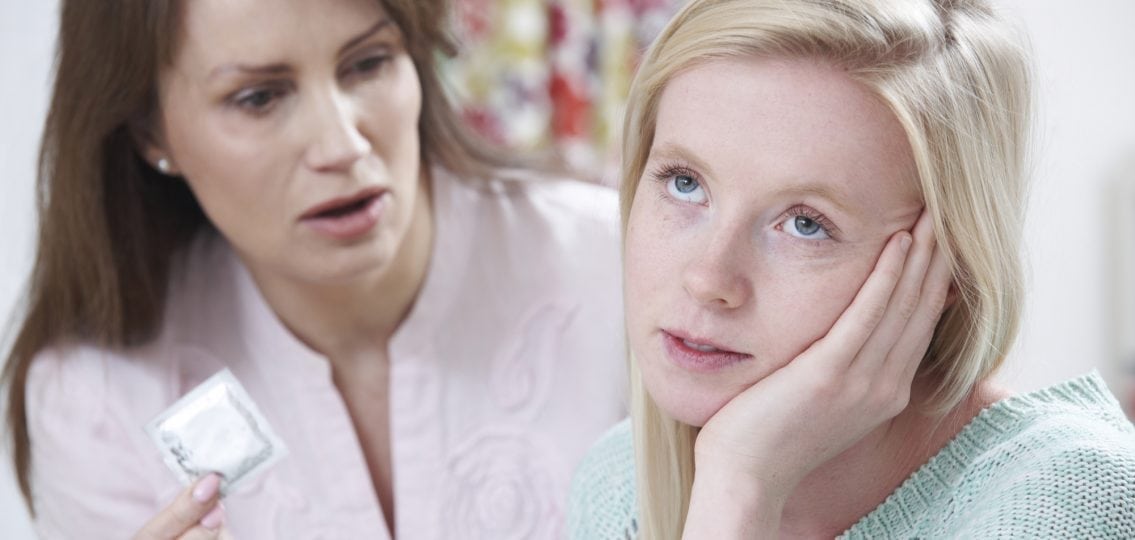 annoyed teen girl rolling her eyes while her distressed mother holds up a condom