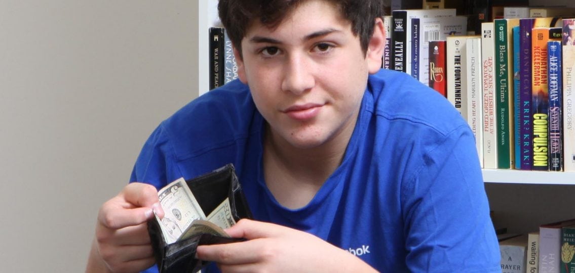 teen boy opening his wallet to show off fifteen dollars in front of a bookshelf