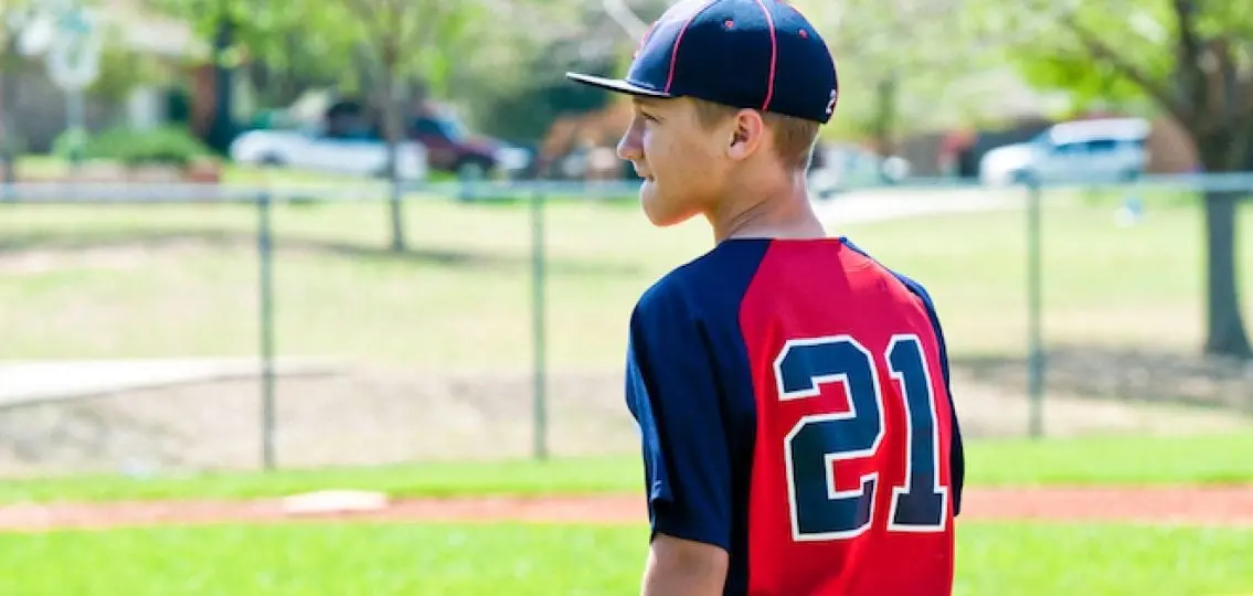 teen boy in baseball uniform biting lip facing away from camera in a baseball field