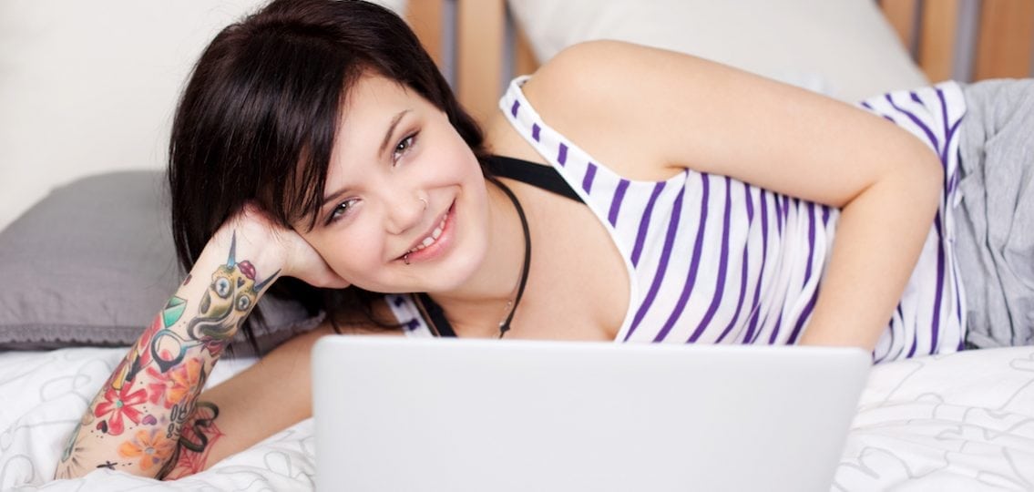 teen girl with sleeve tattoo lying on a bed on her laptop