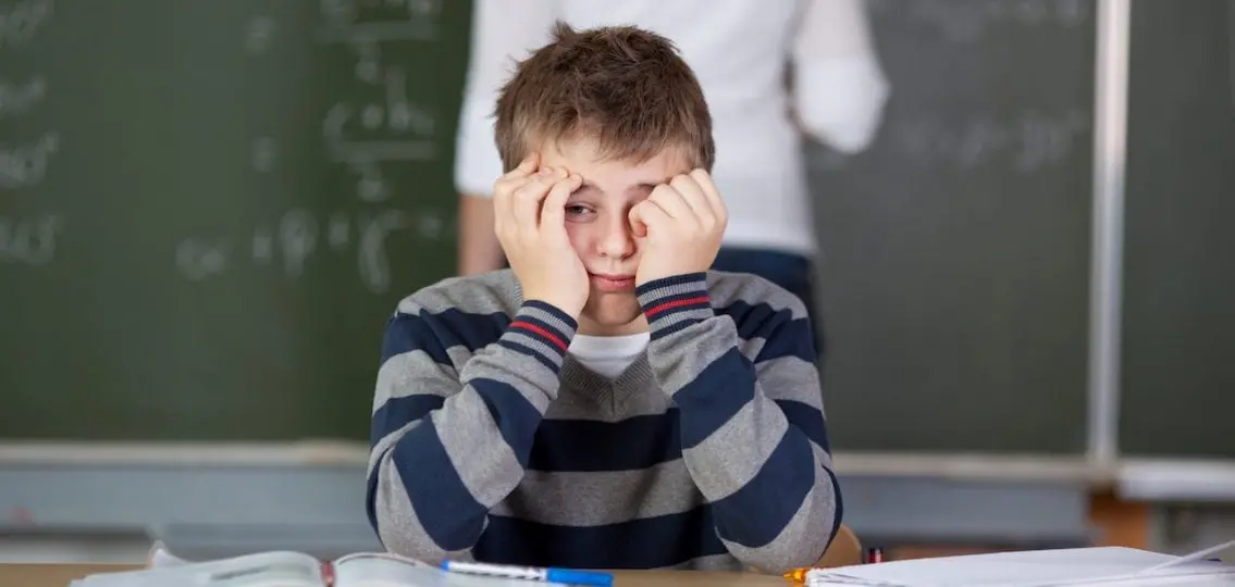 Student With Hands On Face Sitting At Desk while teacher writes on chalkboard in the background
