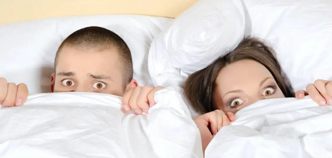 Couple In Bed with covers over their faces eyes wide and horrified
