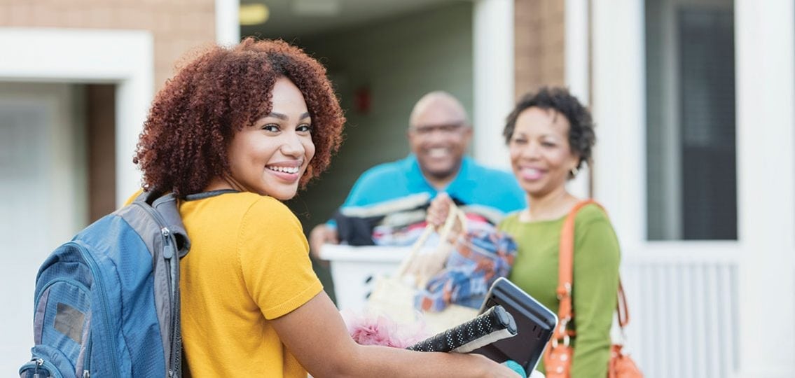 Mature African-American parents helping their daughter relocate, perhaps into an apartment or college dorm. The young woman is in the foreground smiling at the camera, carrying a backpack and basket filled with her belongings.