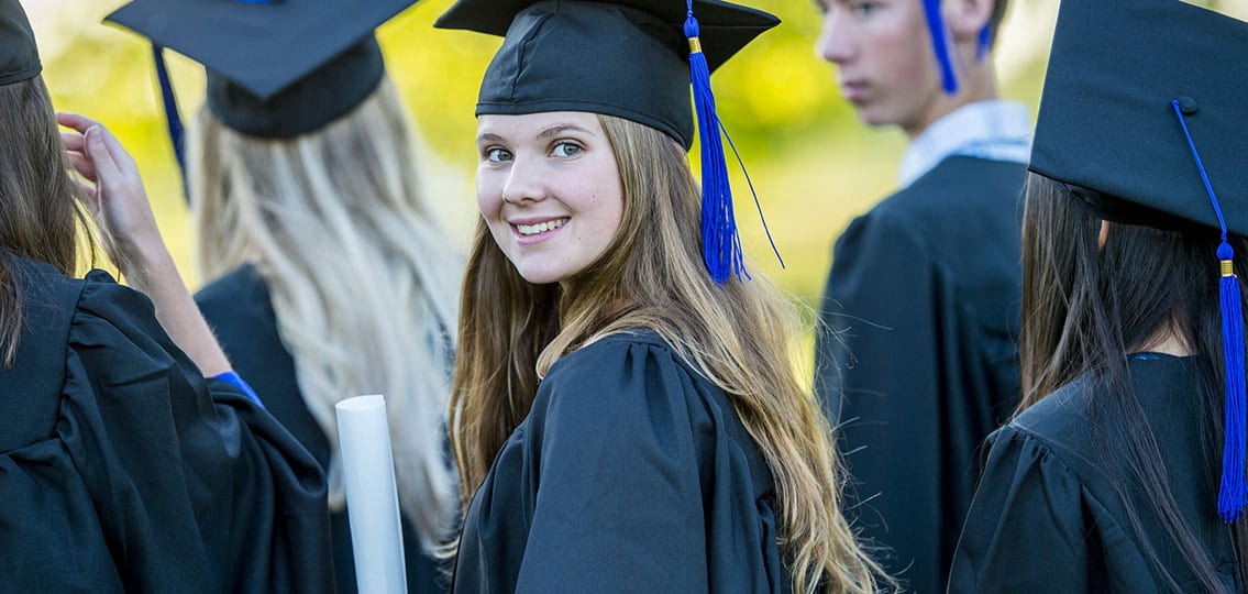 A group of students are at their high school graduation ceremony. They are facing away from the camera, and one girl holding a diploma is turned to smile at the camera.