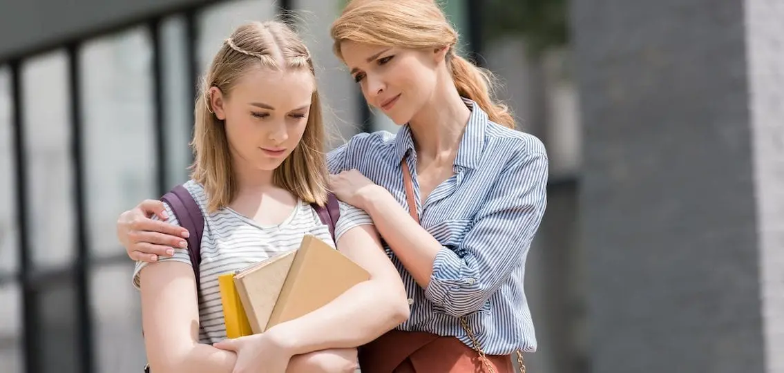 sad mother attempting to comfort sad teen daughter with backpack and school books