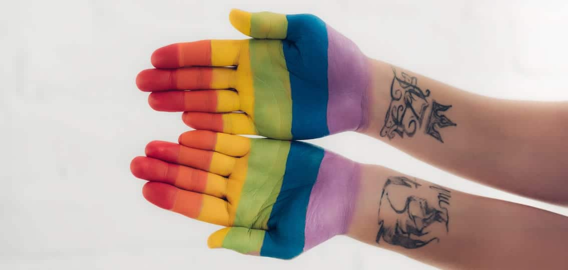 two open hands painted to look like the gay pride rainbow flag with other wrist tattoos